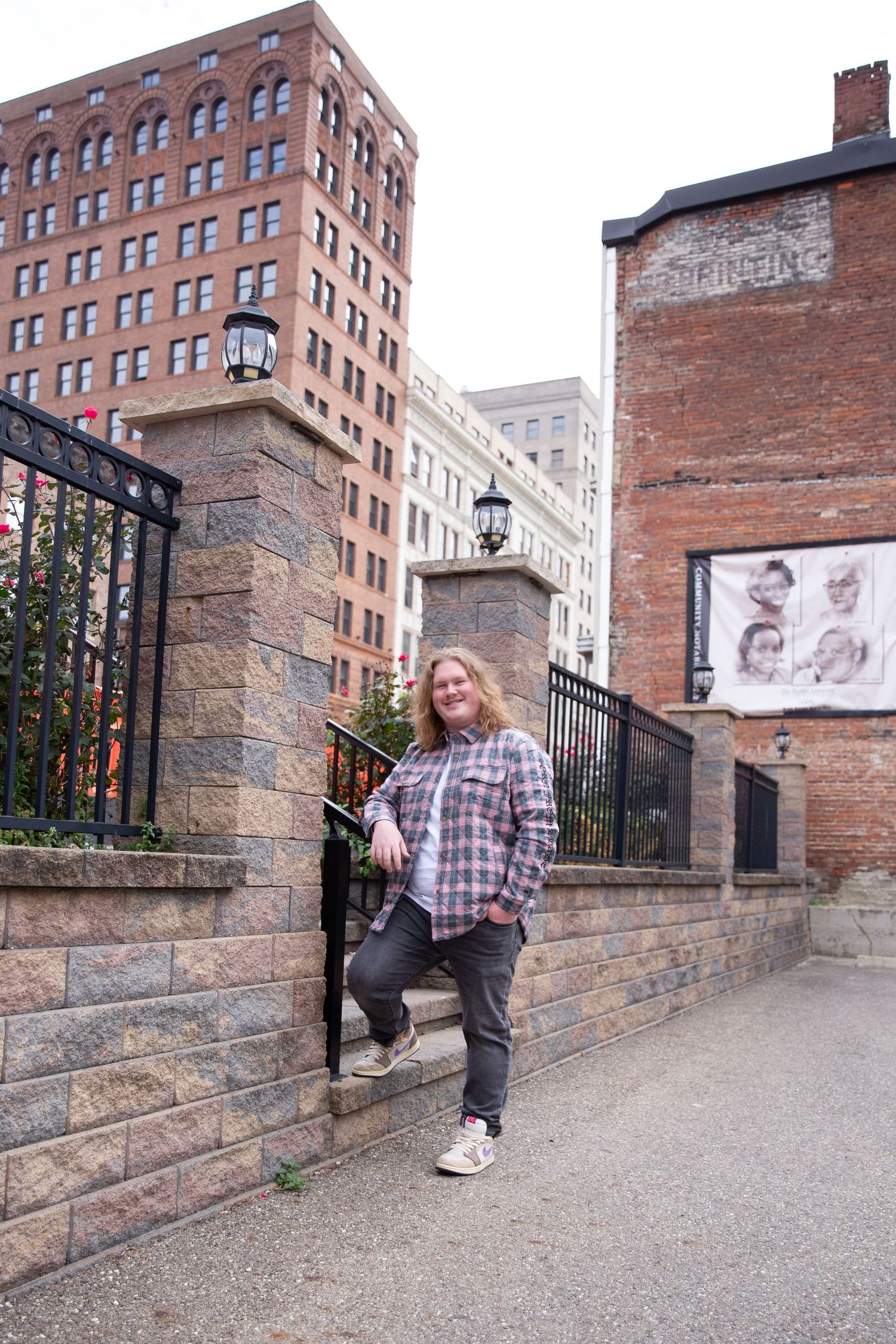 A person poses by historic brick buildings and iron gates in an urban setting.