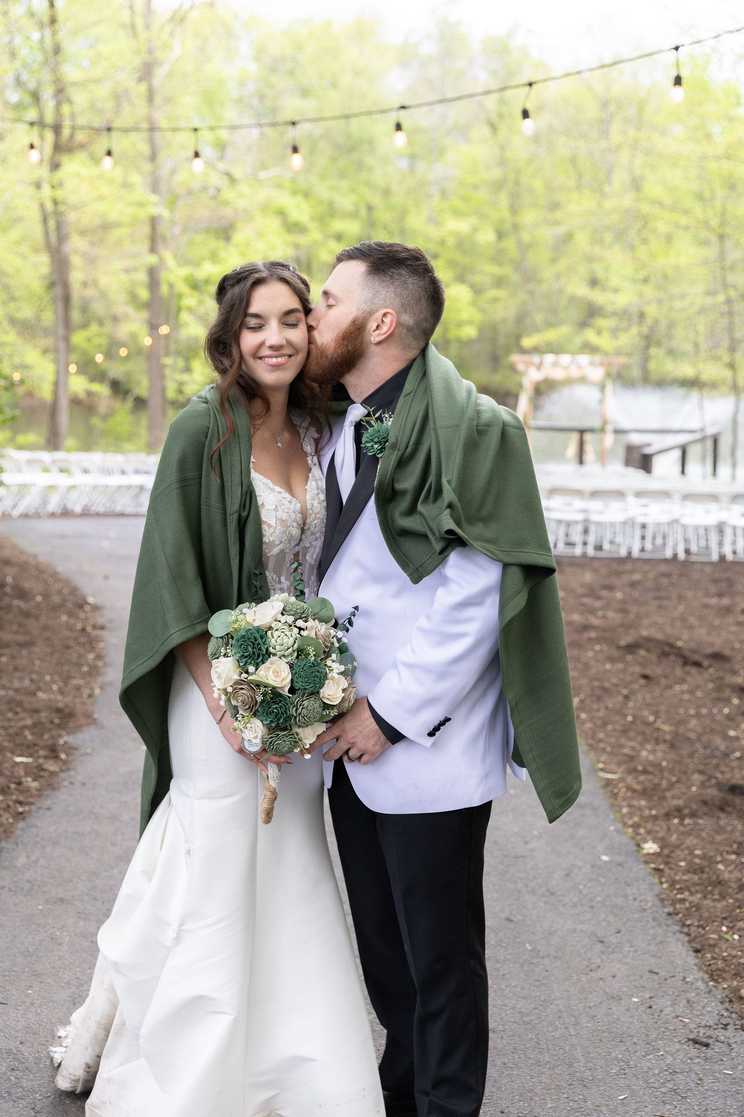 Groom kisses bride on cheek outdoors, bride smiles holding bouquet, both wrapped in green shawl, white wedding attire.