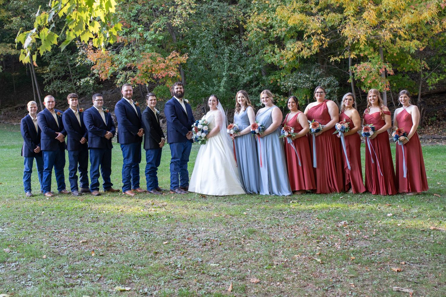 Wedding party lined up outdoors with bridesmaids in burgundy and blue dresses and groomsmen in navy suits.