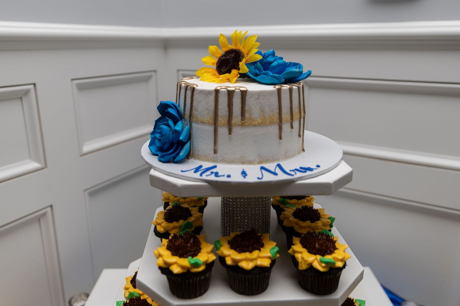 Wedding cake with sunflower and blue floral decor reading Mr. & Mrs., displayed on a tiered stand with matching cupcakes.