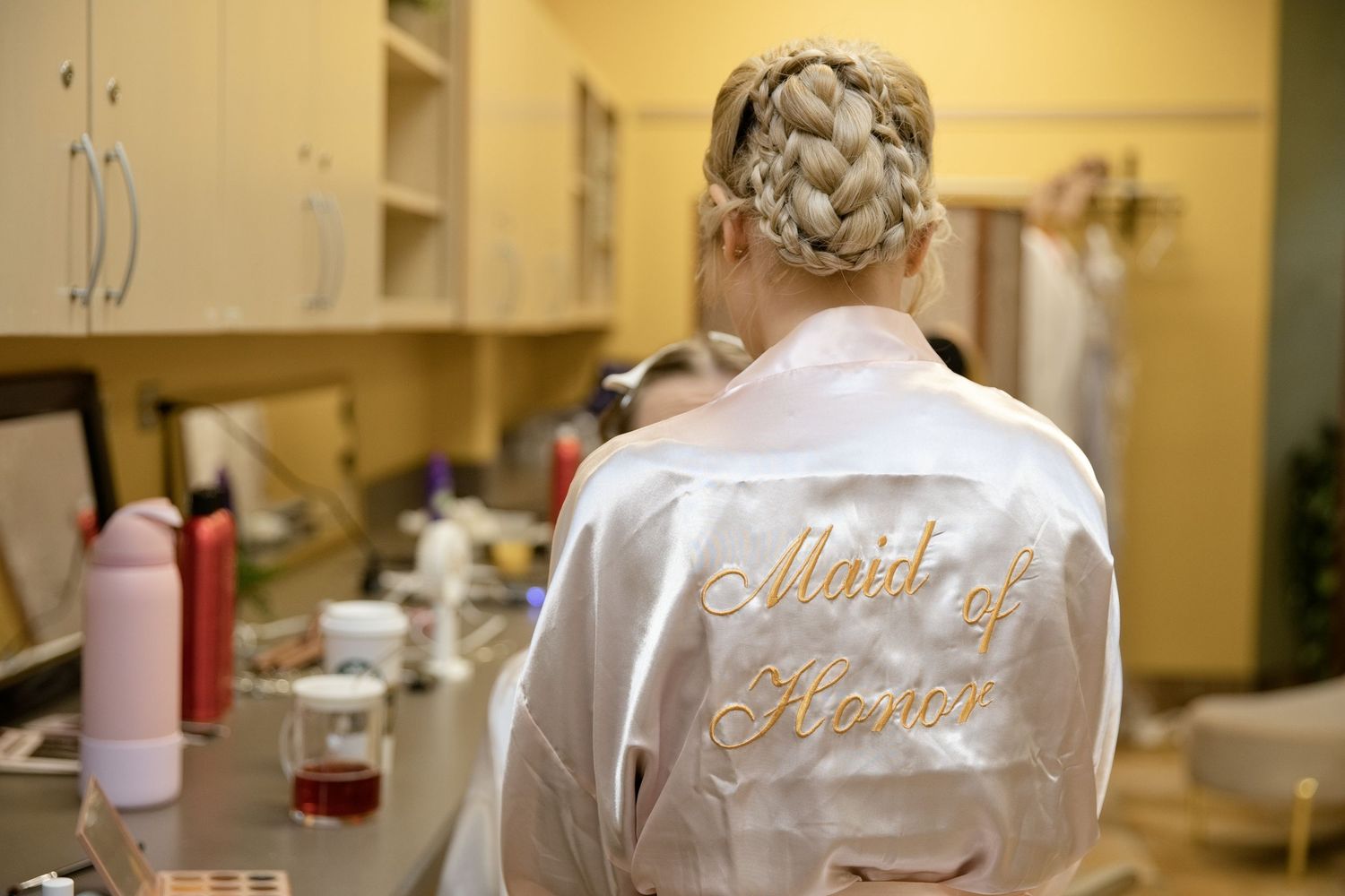 Blonde woman in a white satin Maid of Honor robe with gold embroidery, standing in a bridal preparation room.