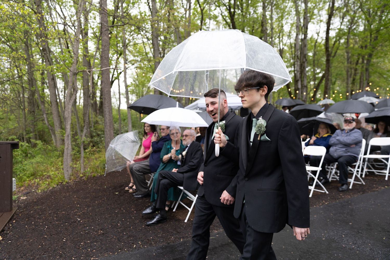 Two grooms walk down the aisle under clear umbrellas at an outdoor rainy wedding ceremony with seated guests.