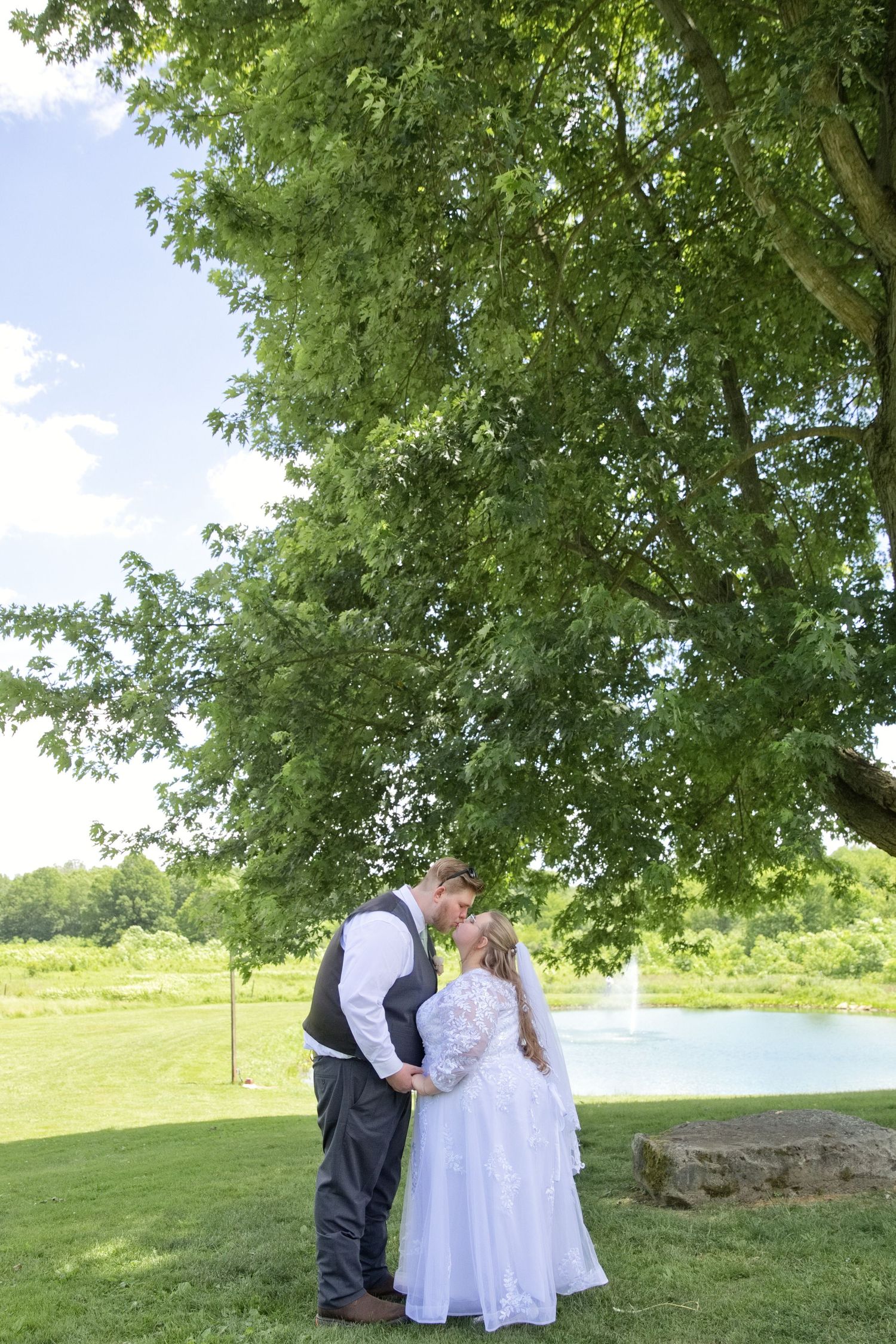 Bride and groom sharing a kiss under a large green tree by a pond on their wedding day.