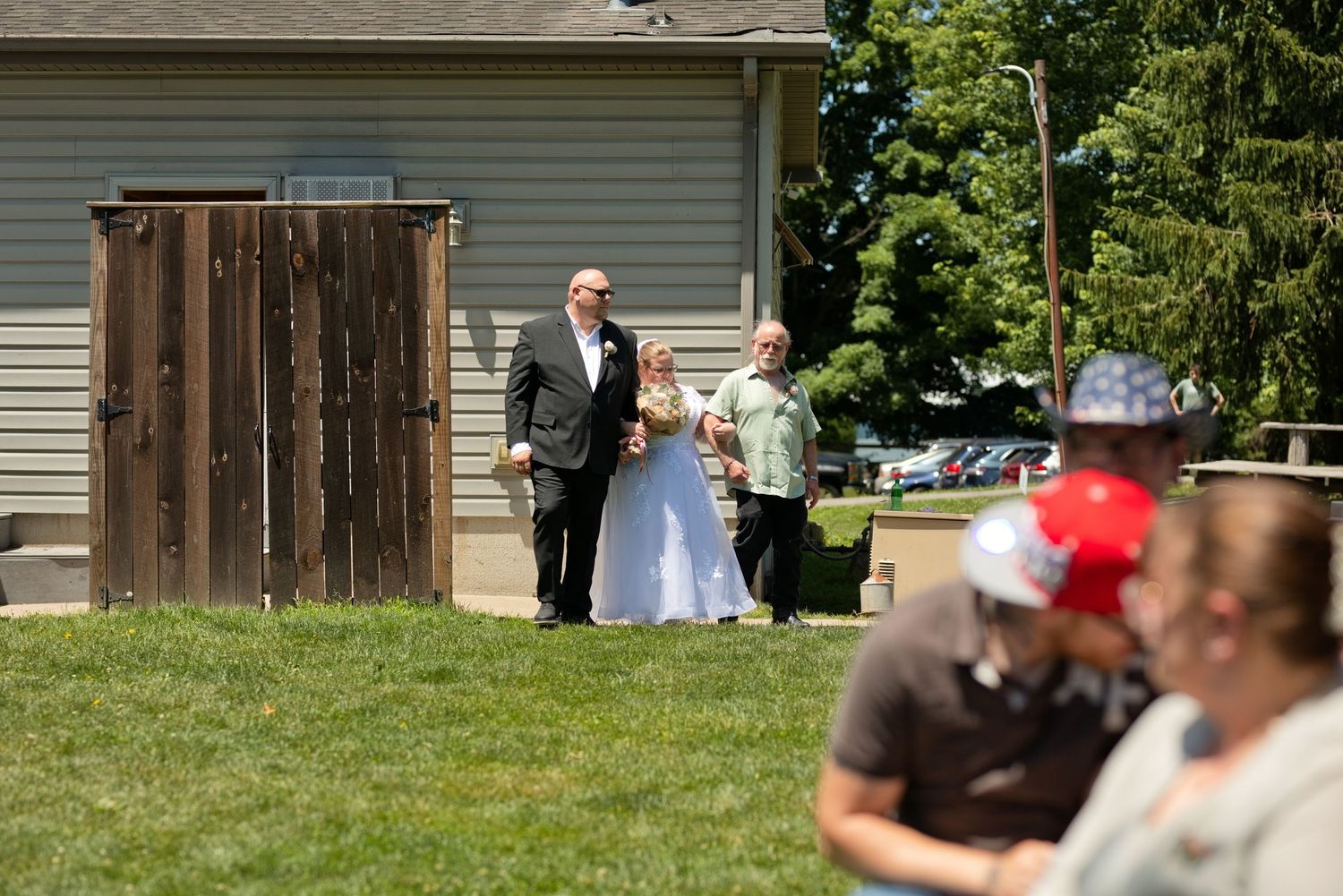Bride and groom walk outdoors at a backyard wedding ceremony on a sunny day with guests in the foreground.