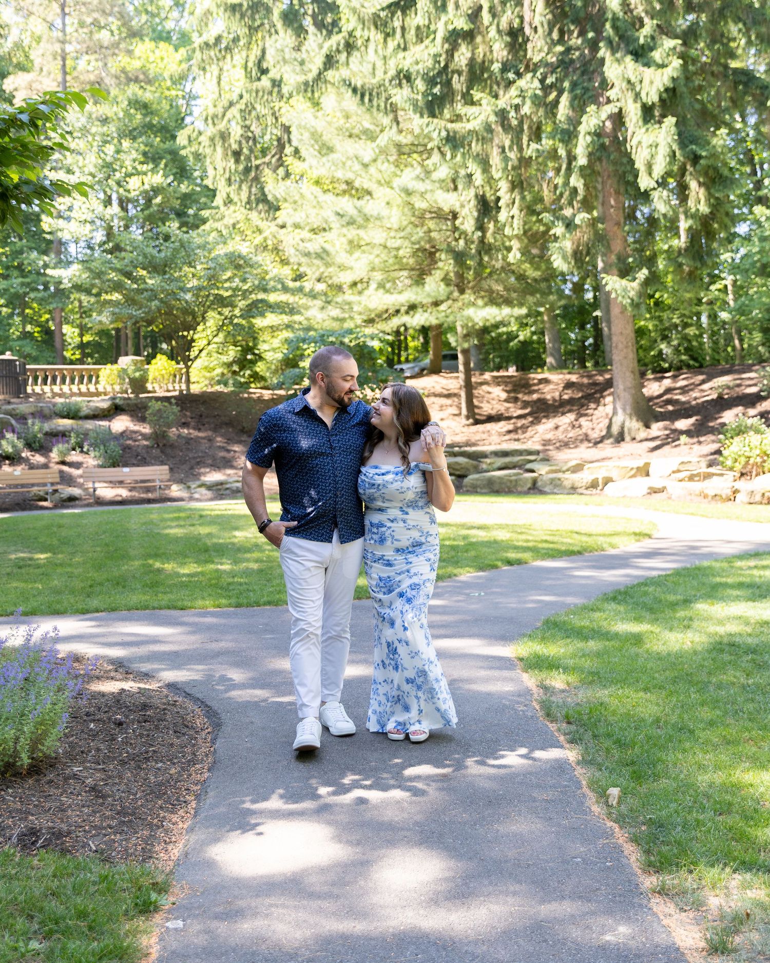 Couple walking together on a garden path surrounded by lush green trees on a sunny day.