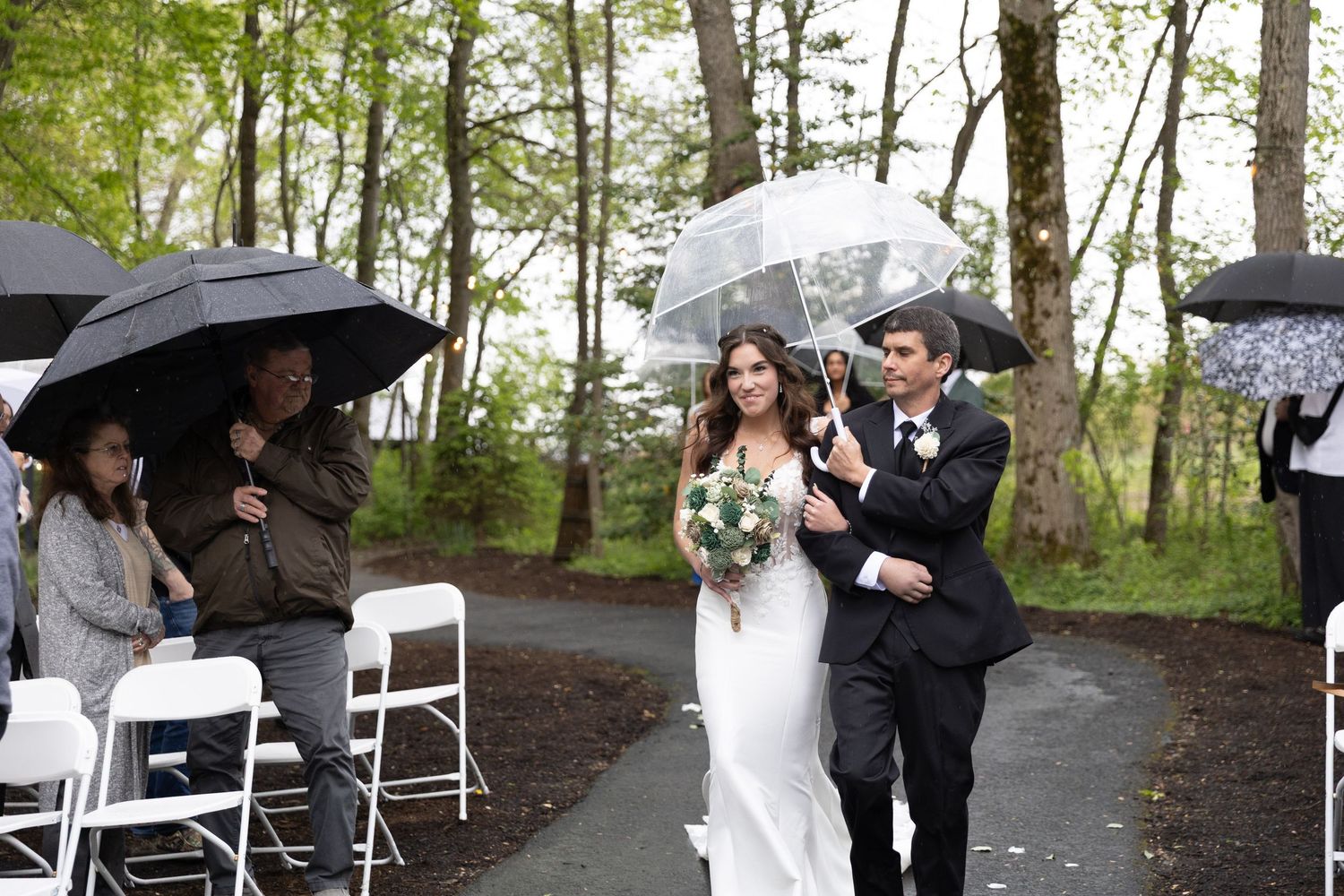 Bride and groom walk down outdoor aisle under clear umbrella during rainy wedding ceremony in wooded setting.