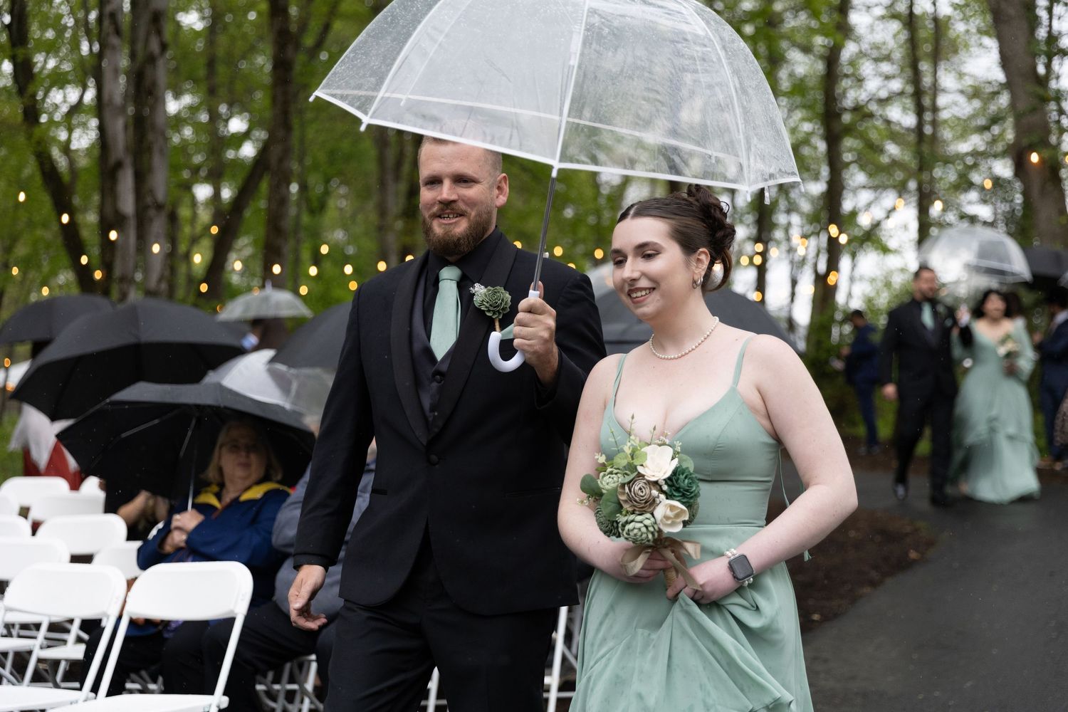 A groomsman and bridesmaid walk down a rainy outdoor wedding aisle sharing a clear umbrella, holding flowers.
