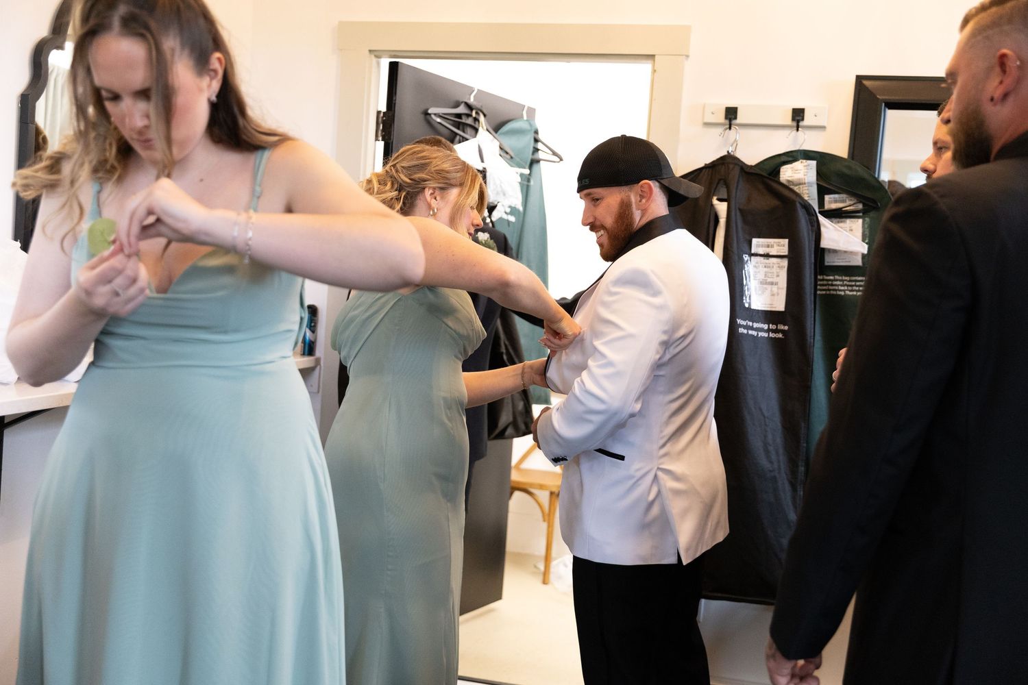 Wedding party getting dressed, bridesmaids in sage green gowns and groomsmen in white suits preparing for ceremony.