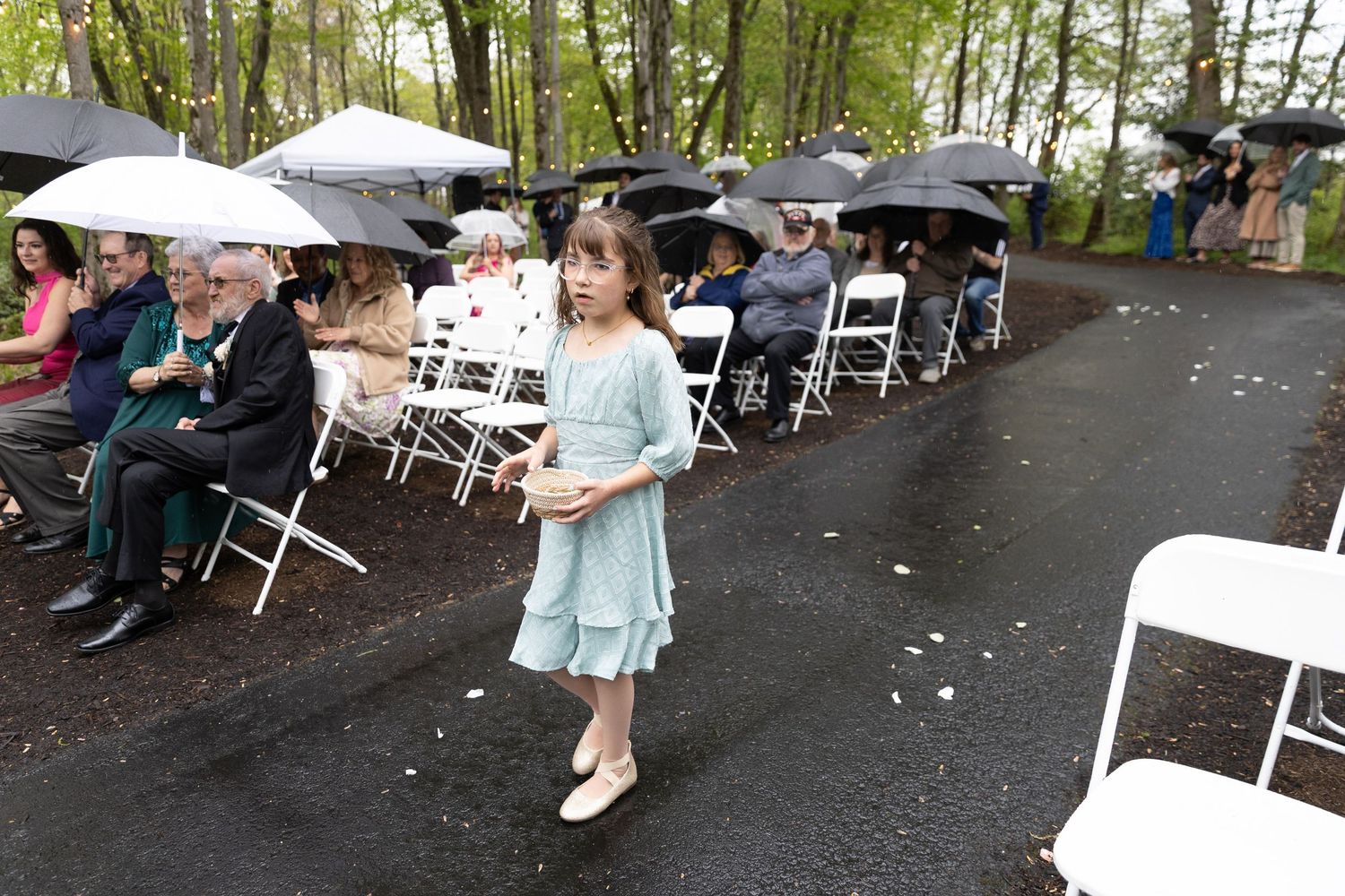 Young flower girl in mint dress walks rainy outdoor wedding aisle while guests sit under umbrellas.