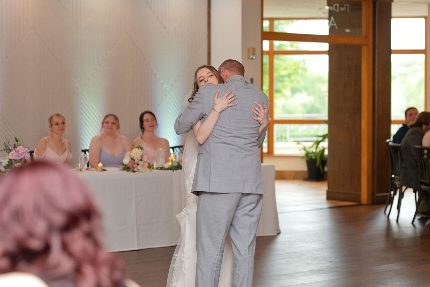 Bride and groom share first dance embrace at wedding reception with bridesmaids seated at head table in background.