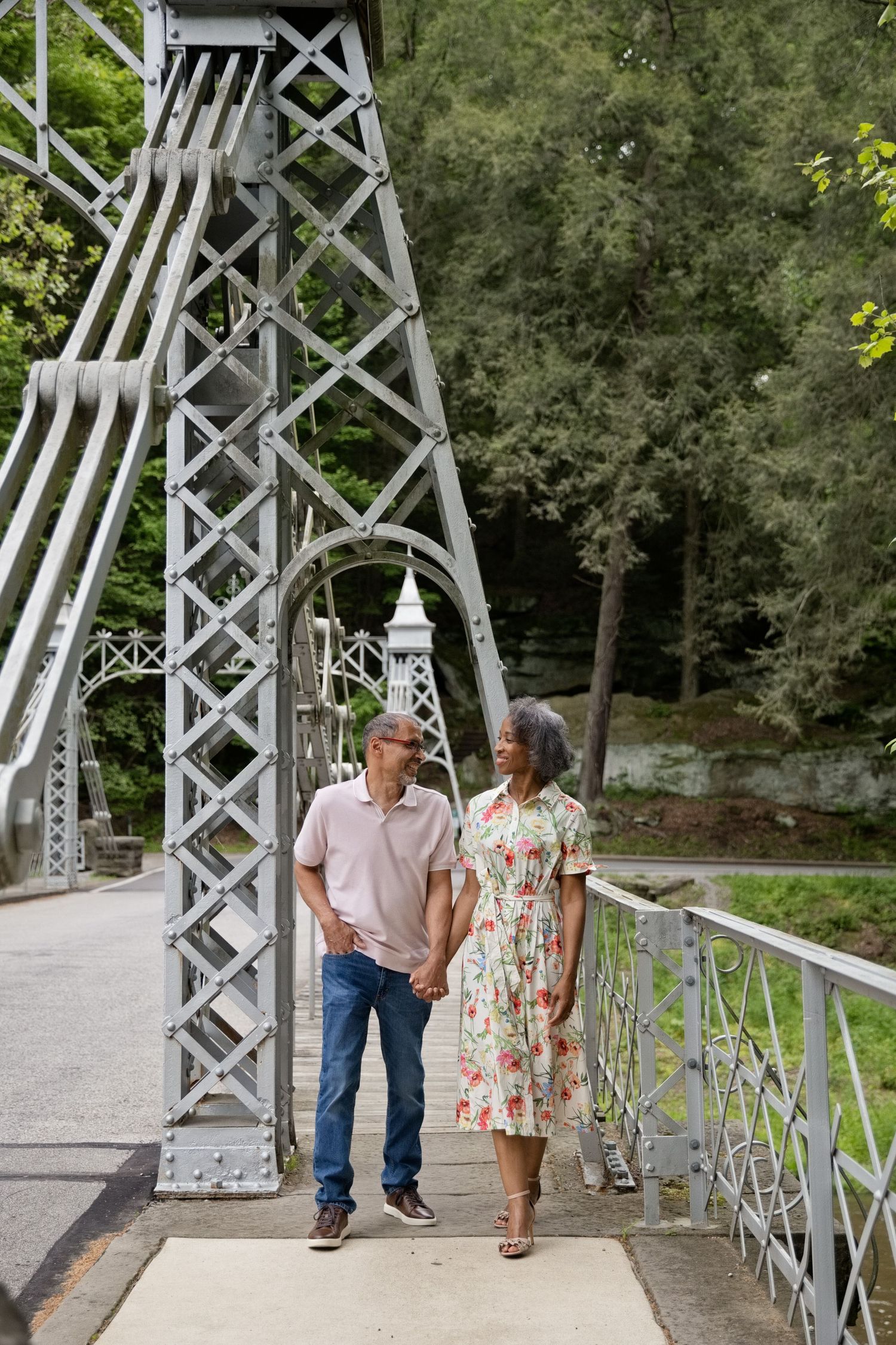 Mature couple holding hands on a white iron bridge surrounded by lush green trees.