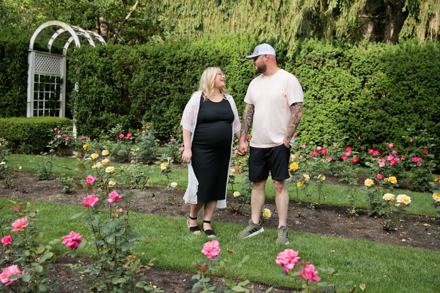 Couple holding hands and walking through a colorful rose garden with lush green hedges and a white arbor.