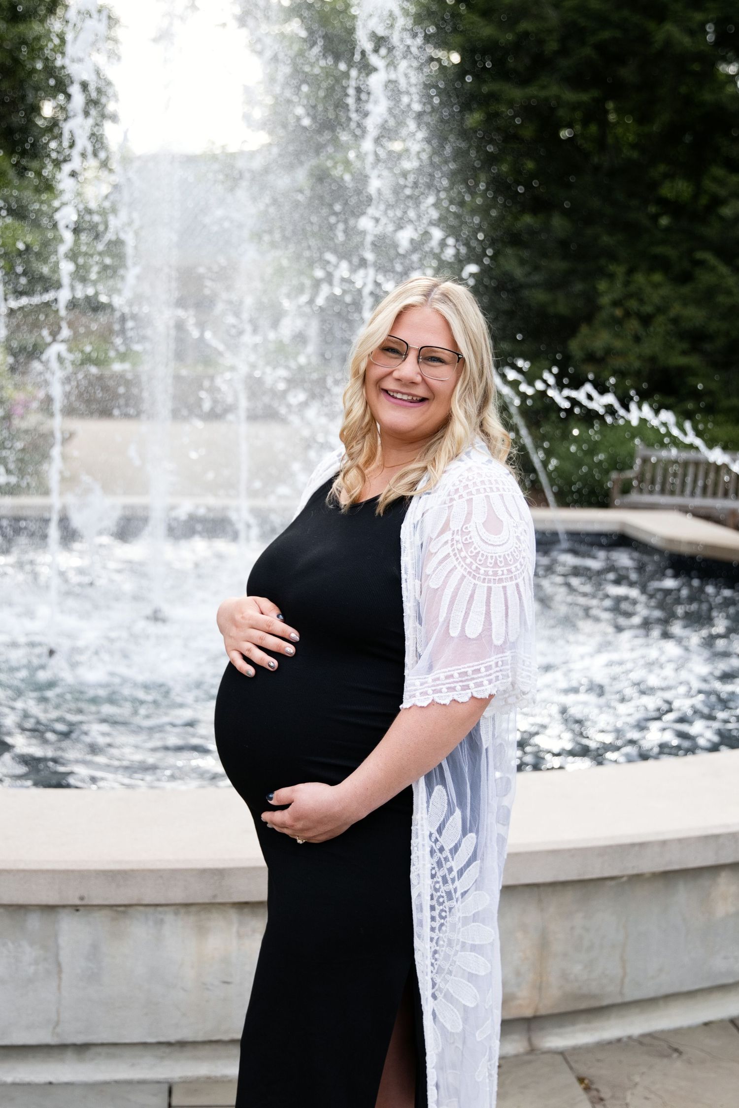 Smiling pregnant woman in black dress and white lace cardigan poses by a fountain outdoors.
