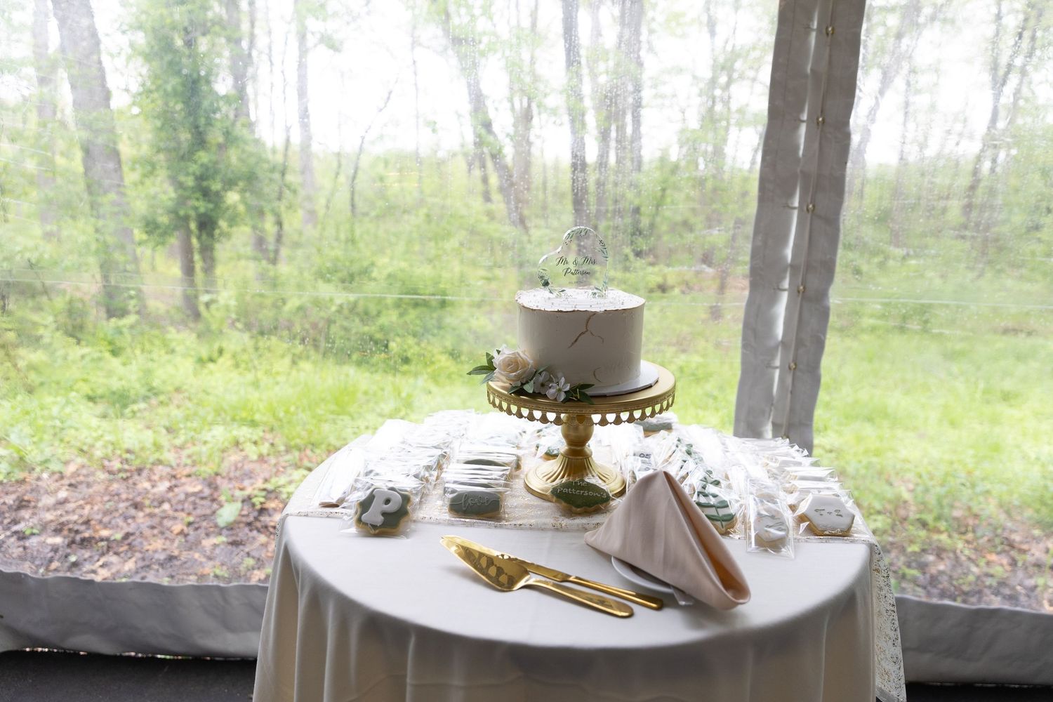 Elegant white wedding cake on gold stand displayed on draped table with forest backdrop.