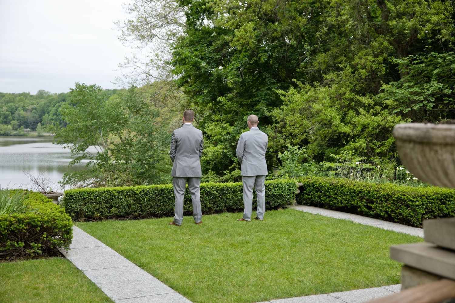 Two men in matching gray suits stand on a manicured lawn overlooking a serene lake surrounded by lush greenery.