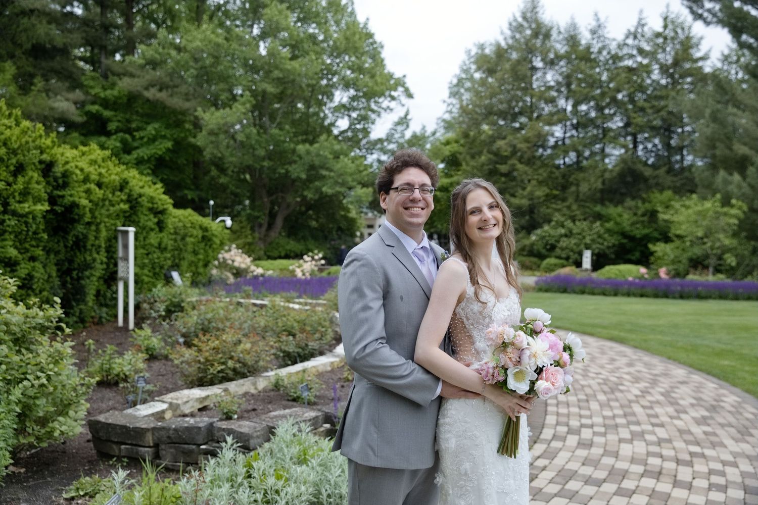 Bride and groom pose in lush garden, bride holding pink bouquet, groom in gray suit.