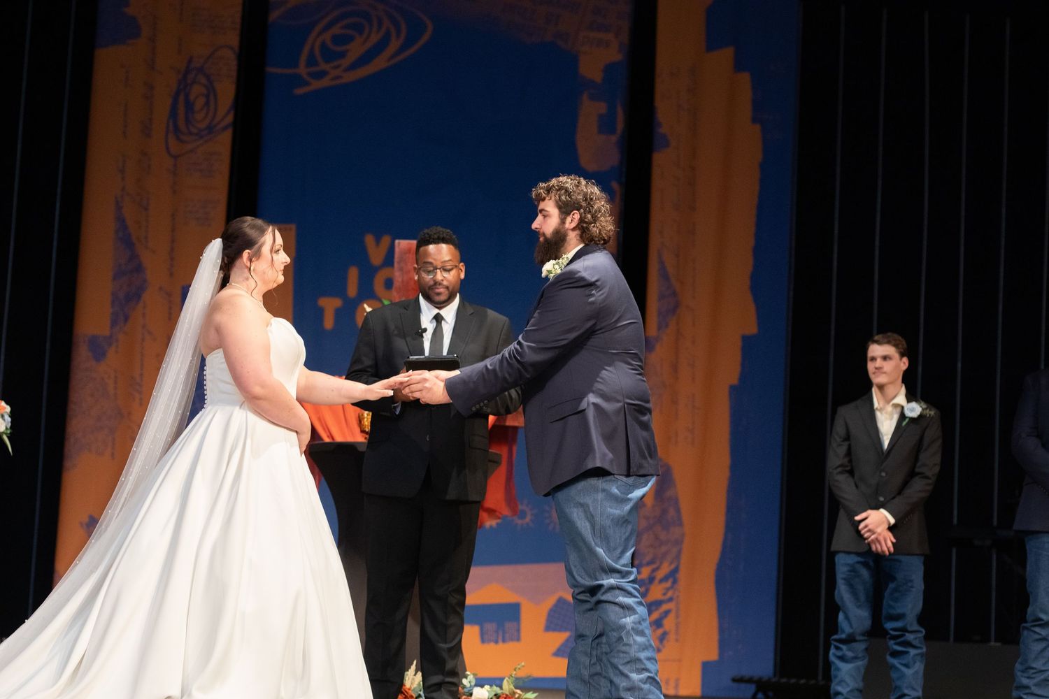 Wedding ceremony exchange of vows on stage with blue backdrop and officiant.