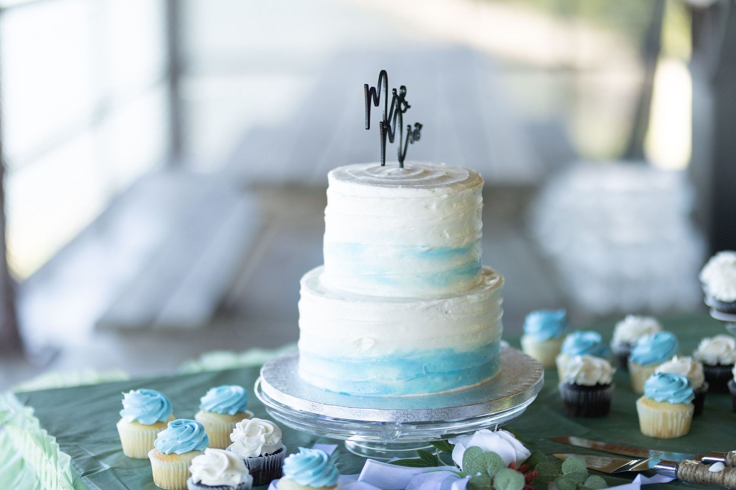 Two-tier wedding cake with blue watercolor design and mini cupcakes on display.