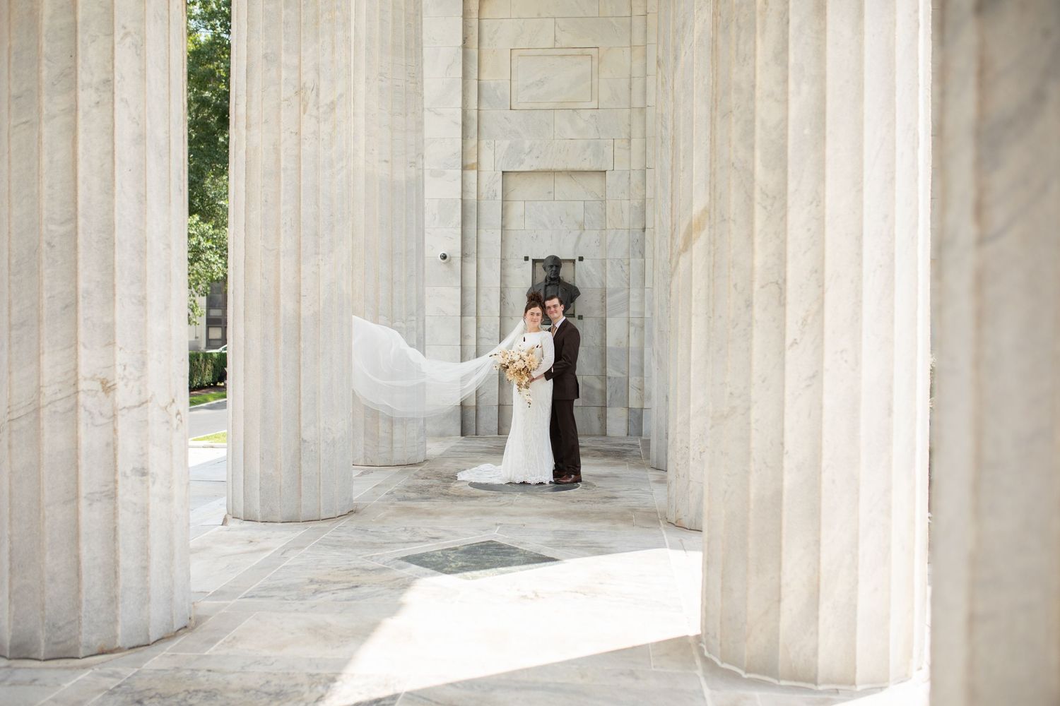 Wedding couple poses between classic white marble columns with flowing veil.