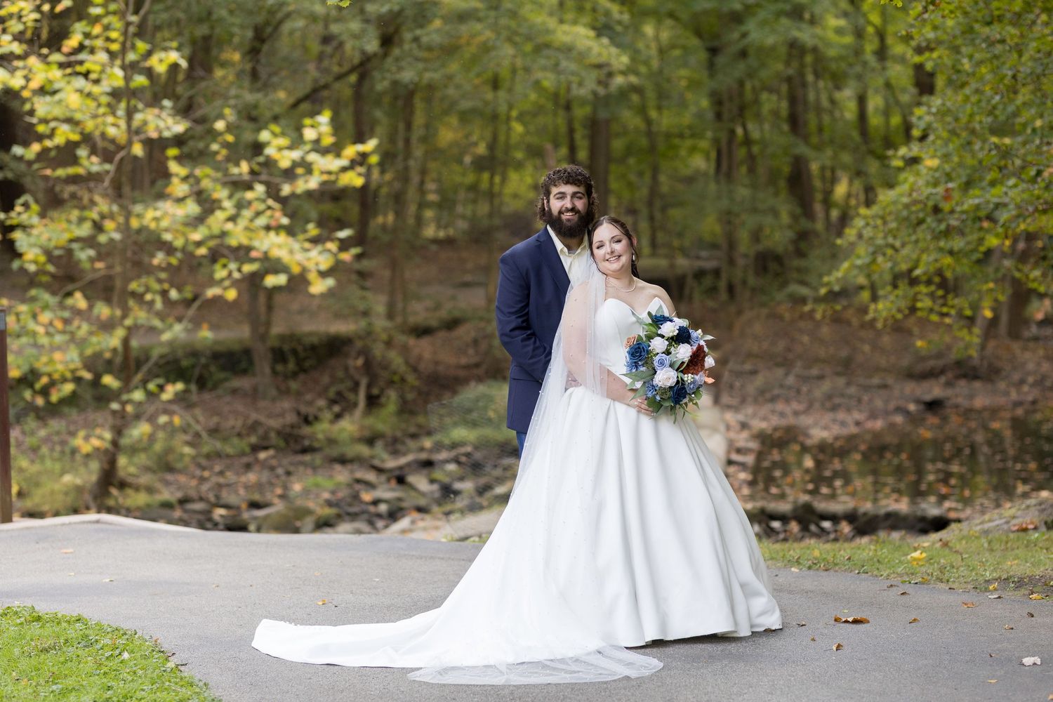 Newlyweds pose together on path with fall foliage in background.
