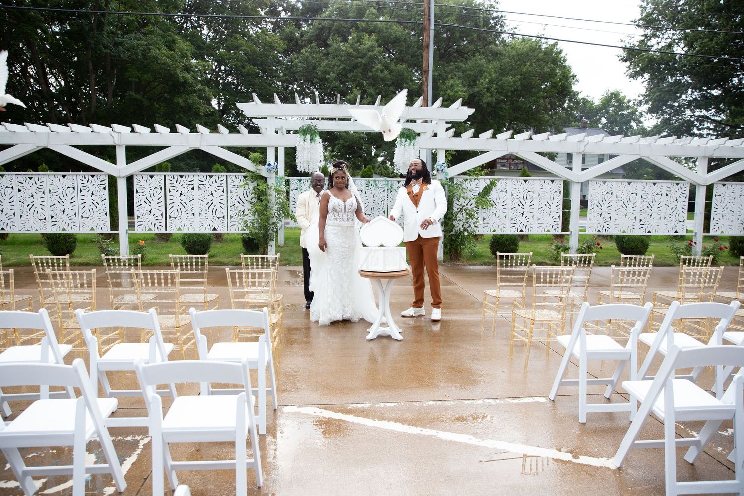 Wedding ceremony setup with white chairs and pergola against decorative wall.