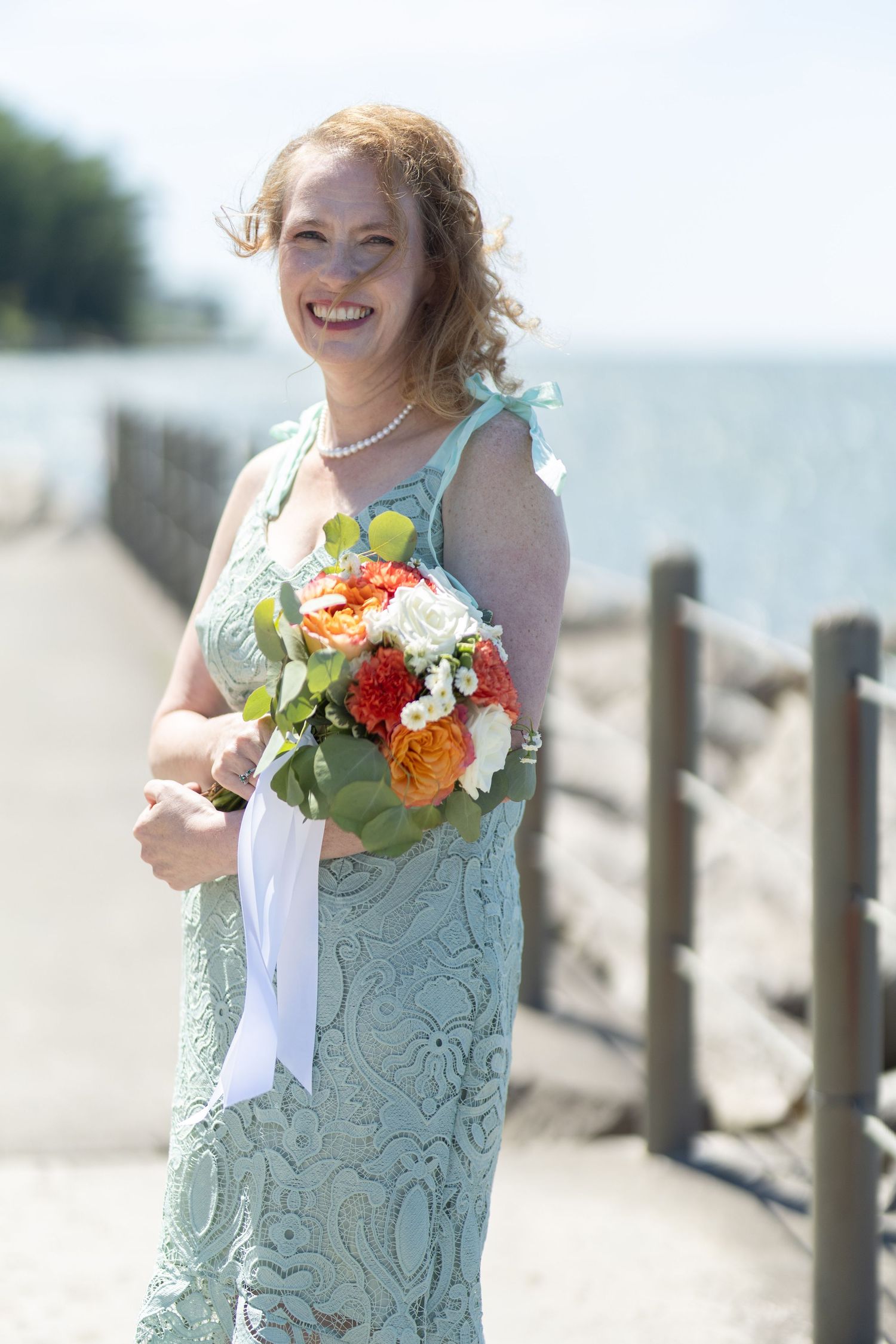 A person in a mint green dress holds a vibrant bouquet of flowers on a sunny beach.