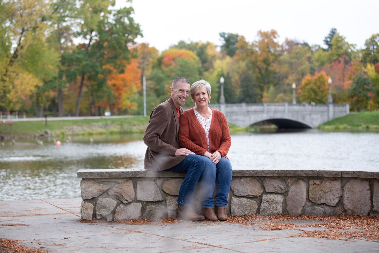 Couple sitting on stone wall surrounded by autumn foliage near a bridge and pond.