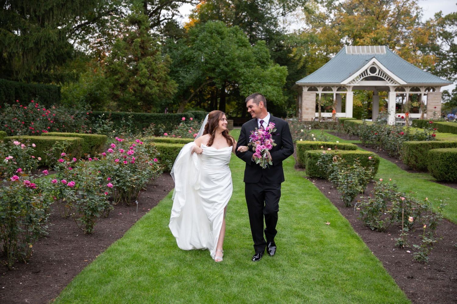 Newlyweds walk through lush garden path with rose bushes and greenery.