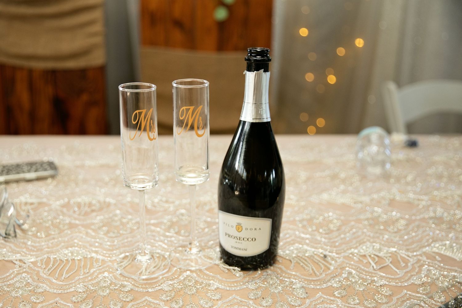 Champagne bottle with two engraved Mr. and Mrs. flute glasses on lace tablecloth at wedding reception.