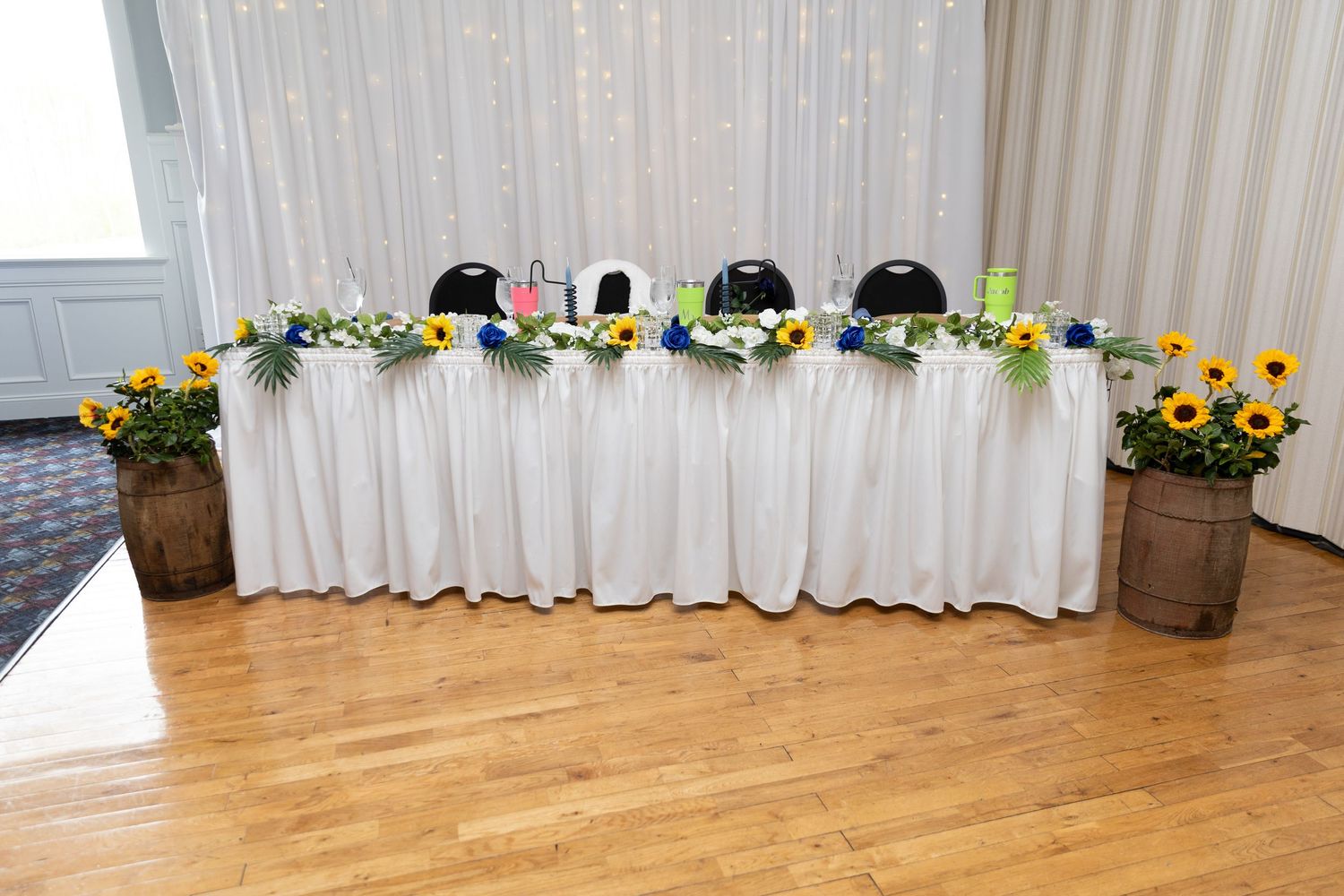 Elegant wedding head table with white linen, sunflower arrangements in wooden barrels, and blue accents in a banquet hall.