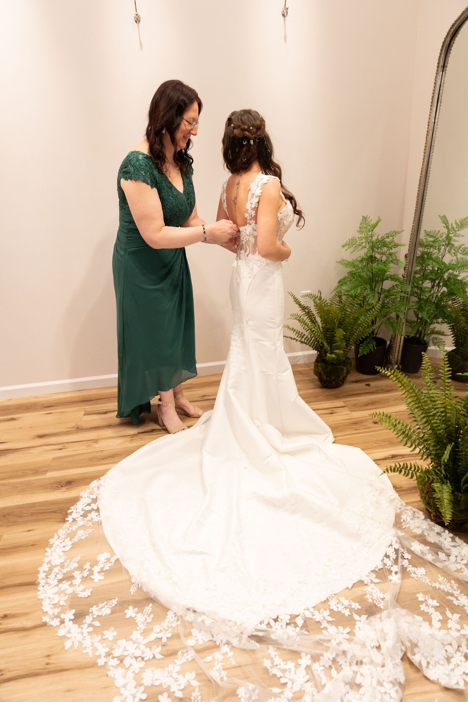 A bride in a white lace gown with train being helped by a woman in green dress in a bright dressing room.