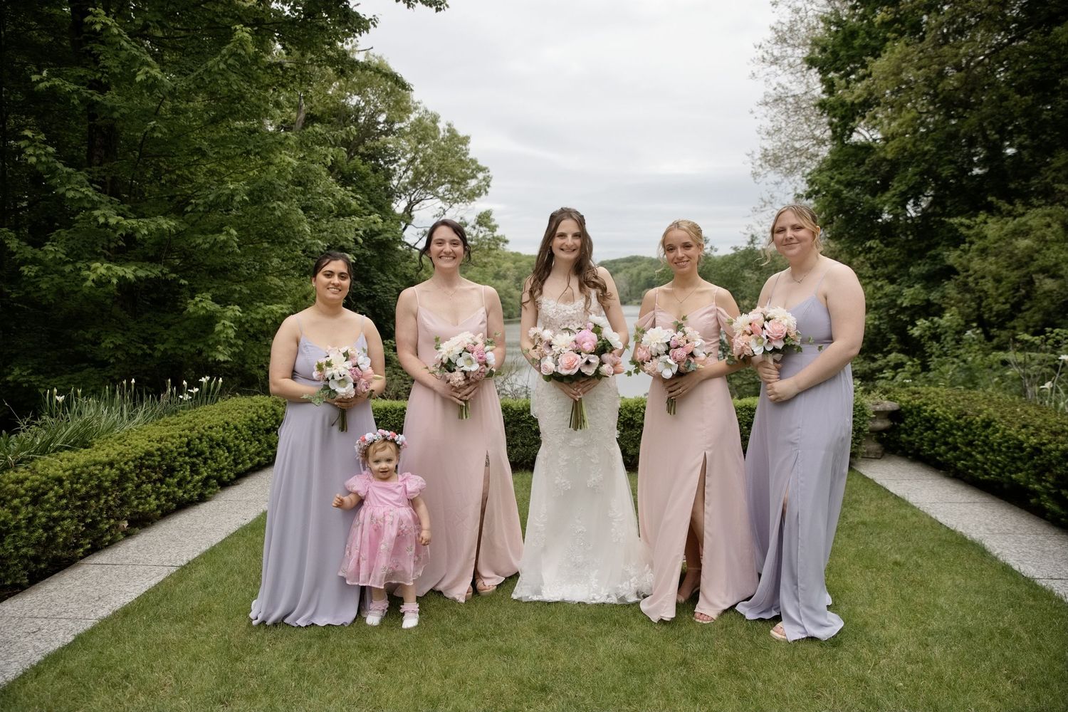 Bride in white gown poses with four bridesmaids in pink and lavender dresses and a flower girl in a garden.