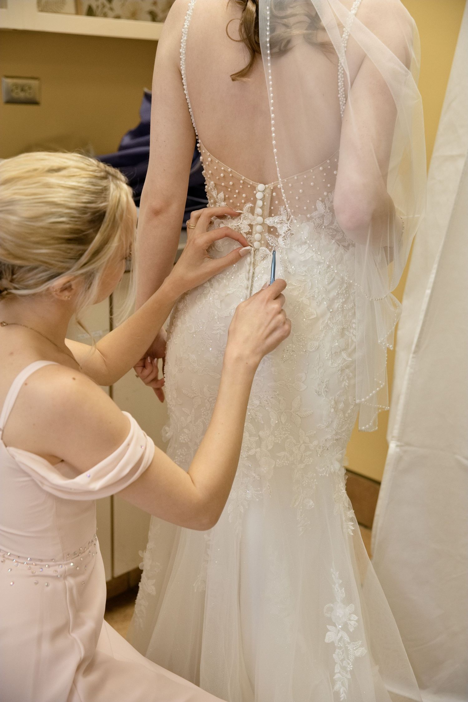 Close-up of bridesmaid fastening pearl buttons on back of brides white lace wedding dress.