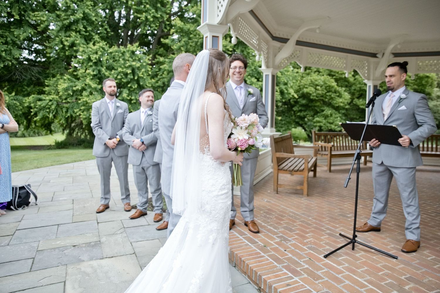 Bride in white gown and veil stands at outdoor gazebo wedding ceremony with groom and groomsmen in gray suits.