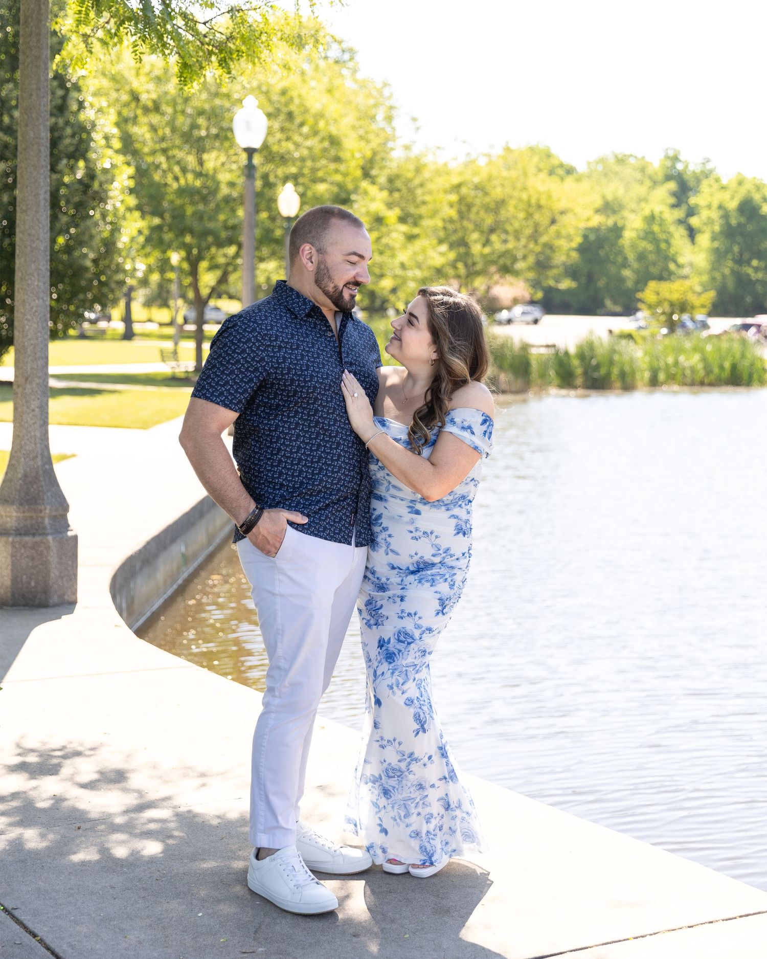 Smiling couple gazing at each other near a lakeside lamppost on a bright summer day.