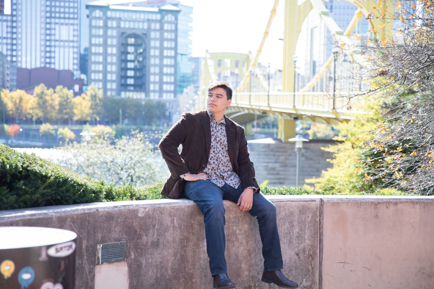 Young man in blazer sits on stone wall with Pittsburgh bridge and skyline backdrop.