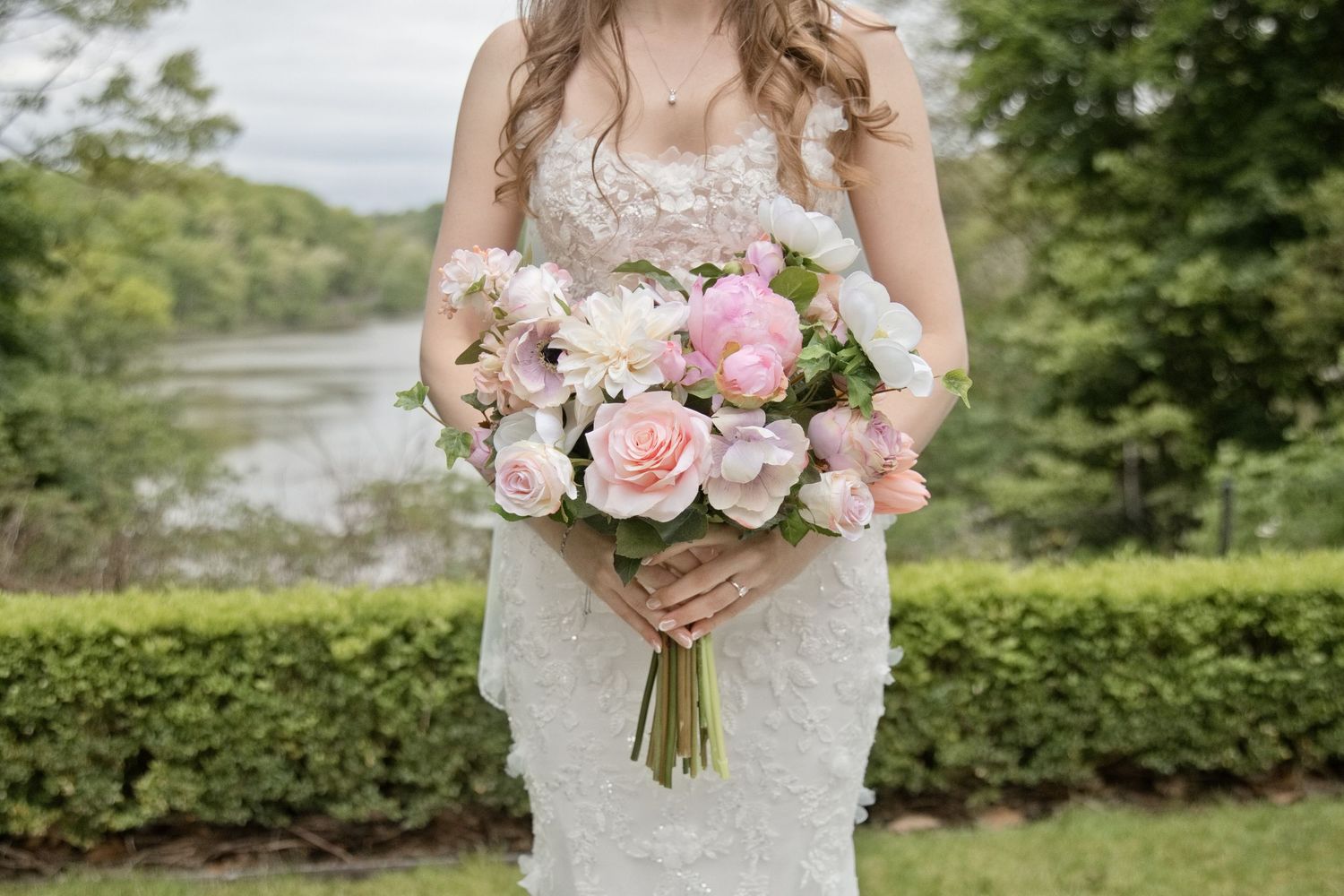 Bride in lace wedding gown holding a pink and white floral bouquet with roses and peonies outdoors near a pond.