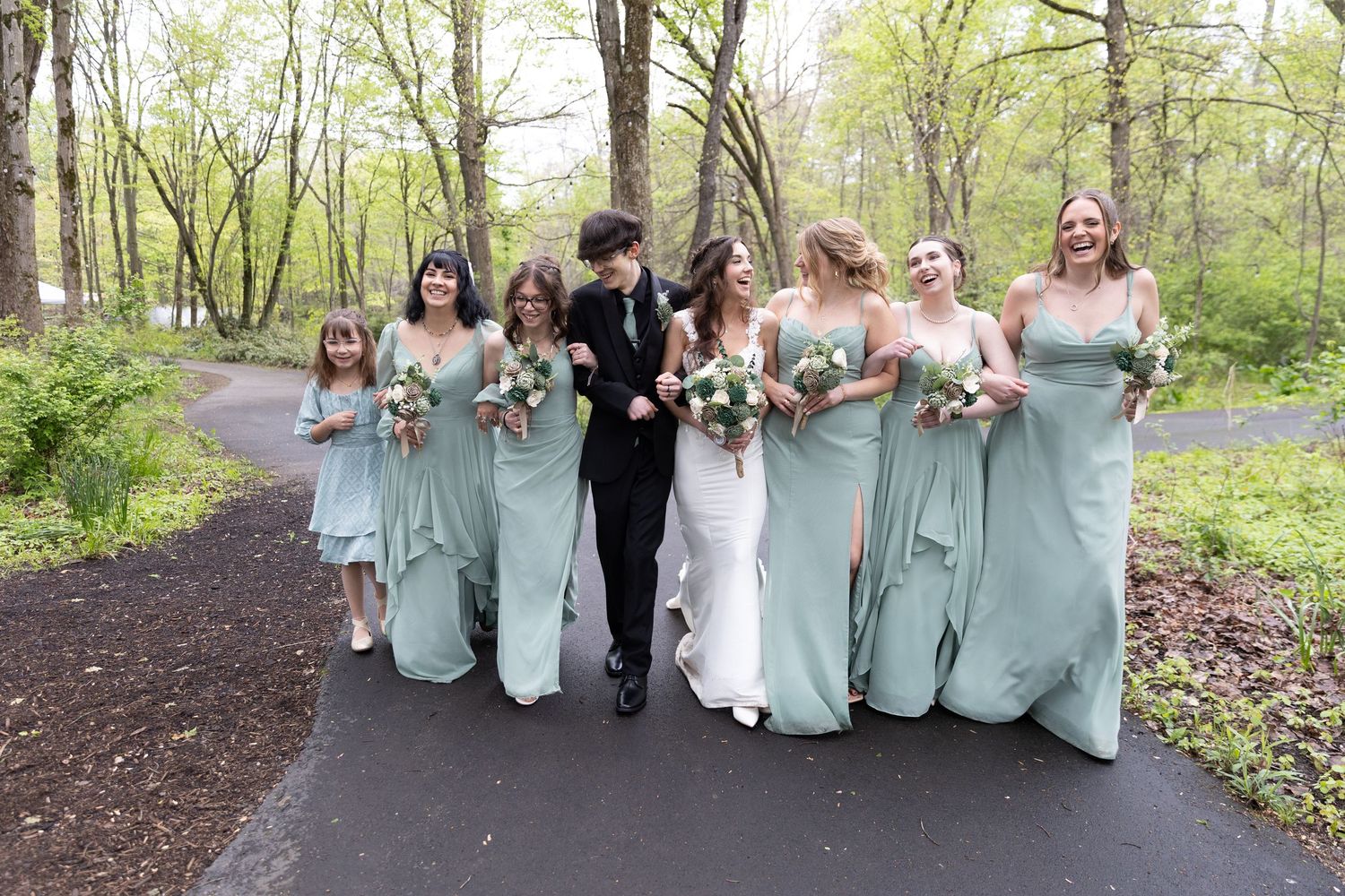 Bride and groom walk with bridesmaids in sage green dresses along a tree-lined path at an outdoor wedding.