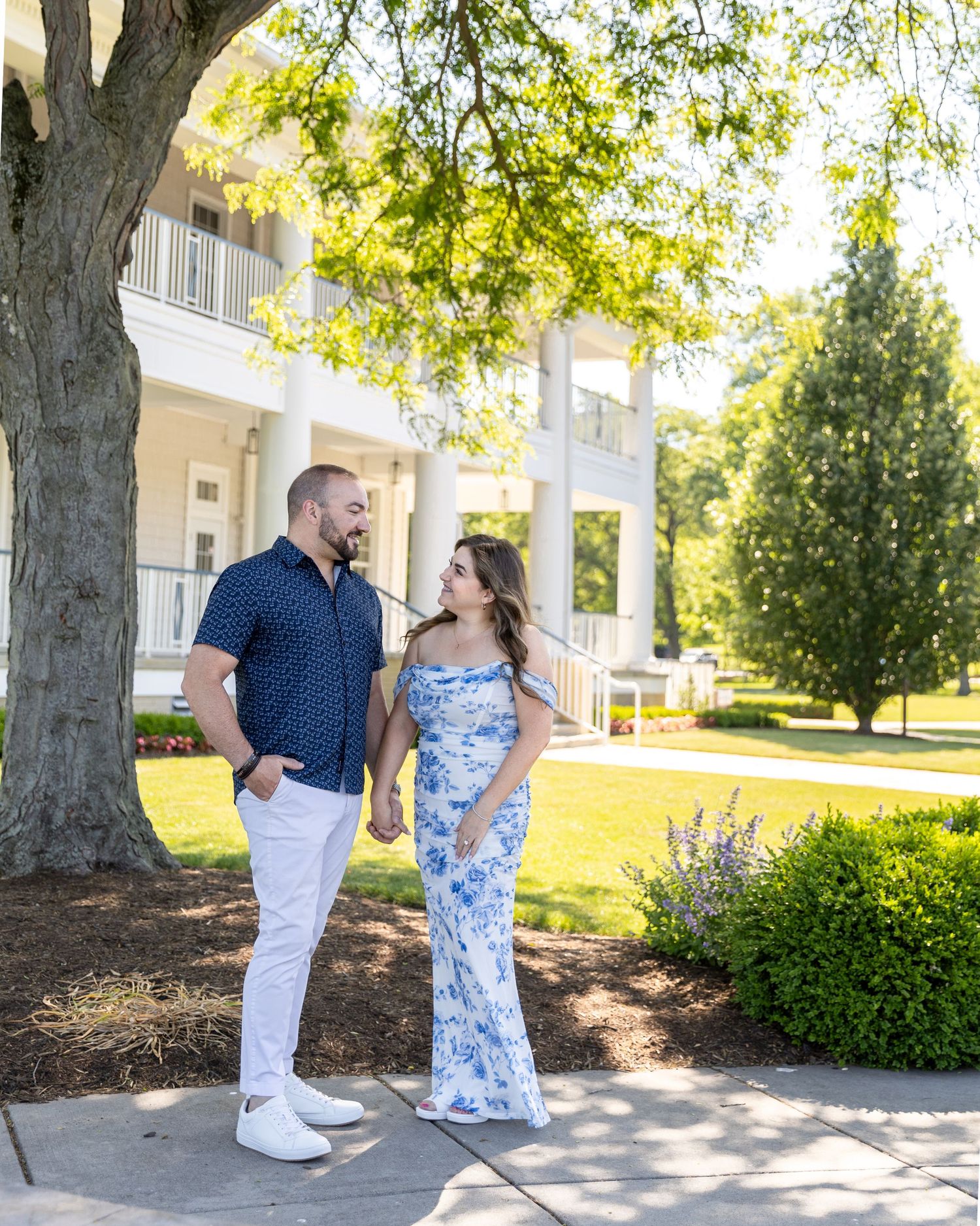 Couple holding hands and gazing at each other on a sunny day outside a white colonial building with lush greenery.