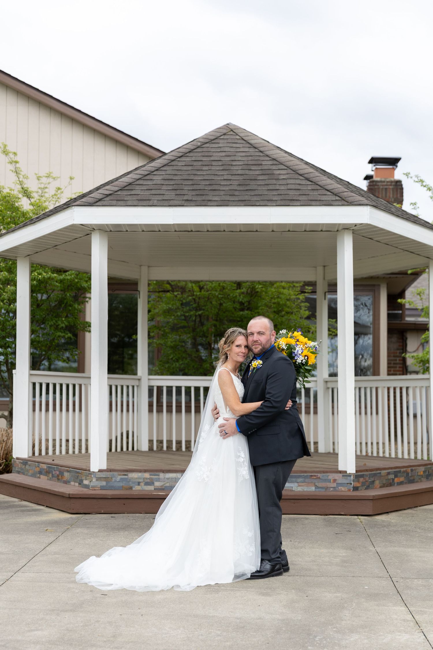 Bride and groom posing in front of a white gazebo, bride in flowing gown holding yellow bouquet on wedding day.