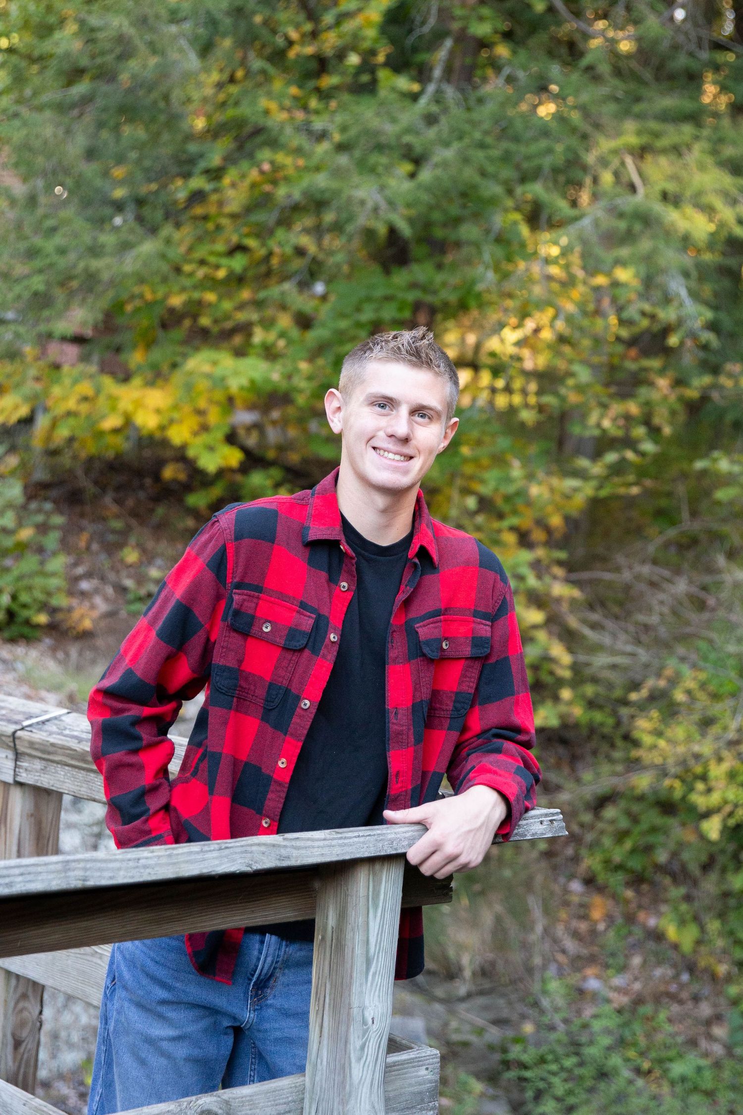A person in a red and black buffalo plaid jacket stands on a wooden deck railing with autumn foliage in background.