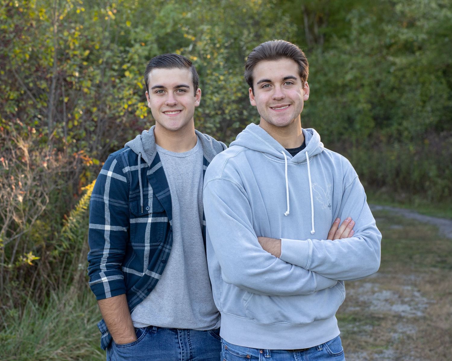 Two young men smiling outdoors, one in a plaid shirt and the other in a gray hoodie, standing on a path with trees behind them.