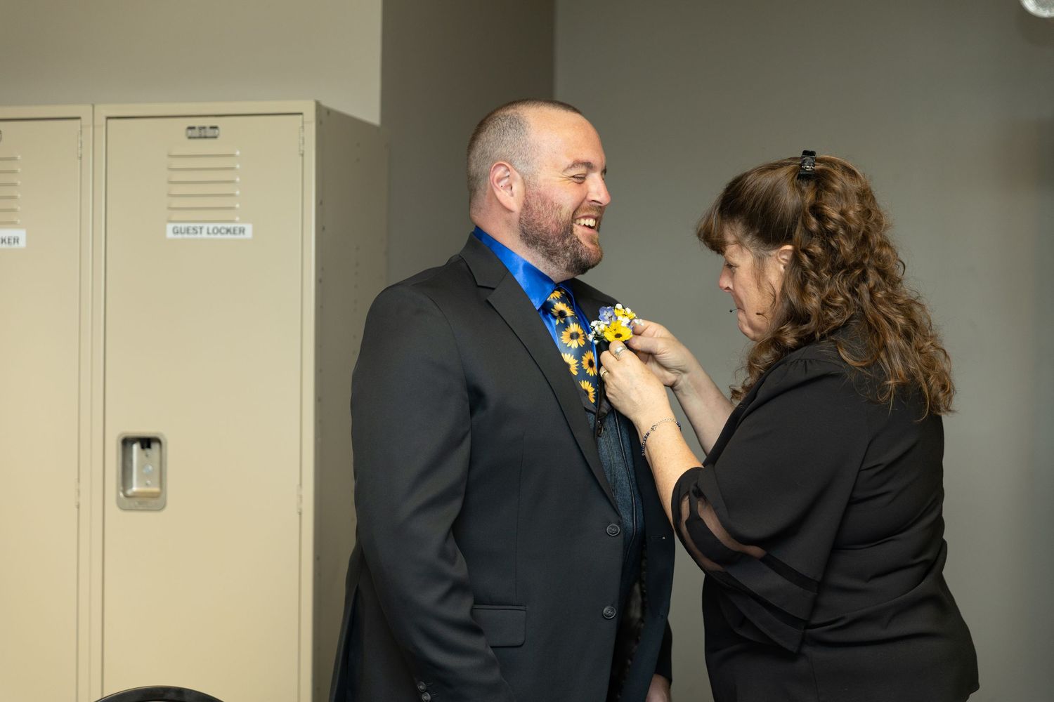 Woman pinning a boutonniere on a man in a dark suit near school lockers.