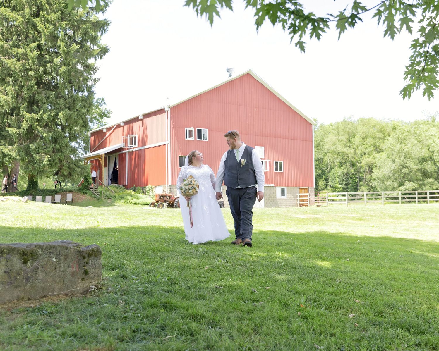 Newlywed couple holding hands walking on green lawn beside a red barn on a sunny wedding day.