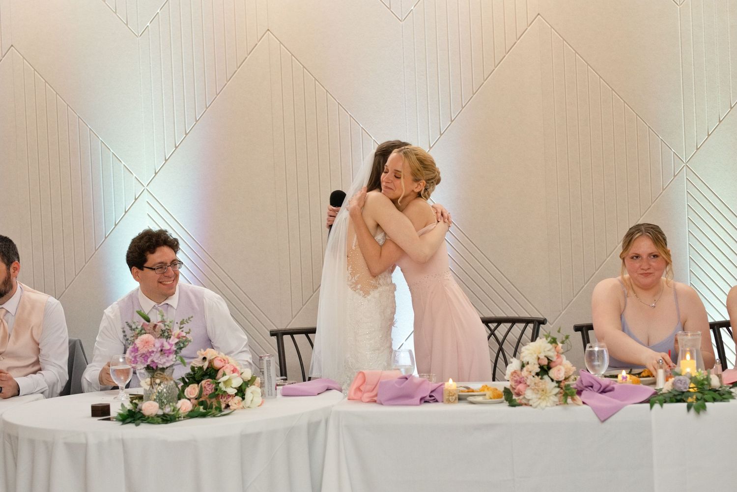 Two women share a heartfelt hug at a wedding reception while guests watch at a decorated head table.