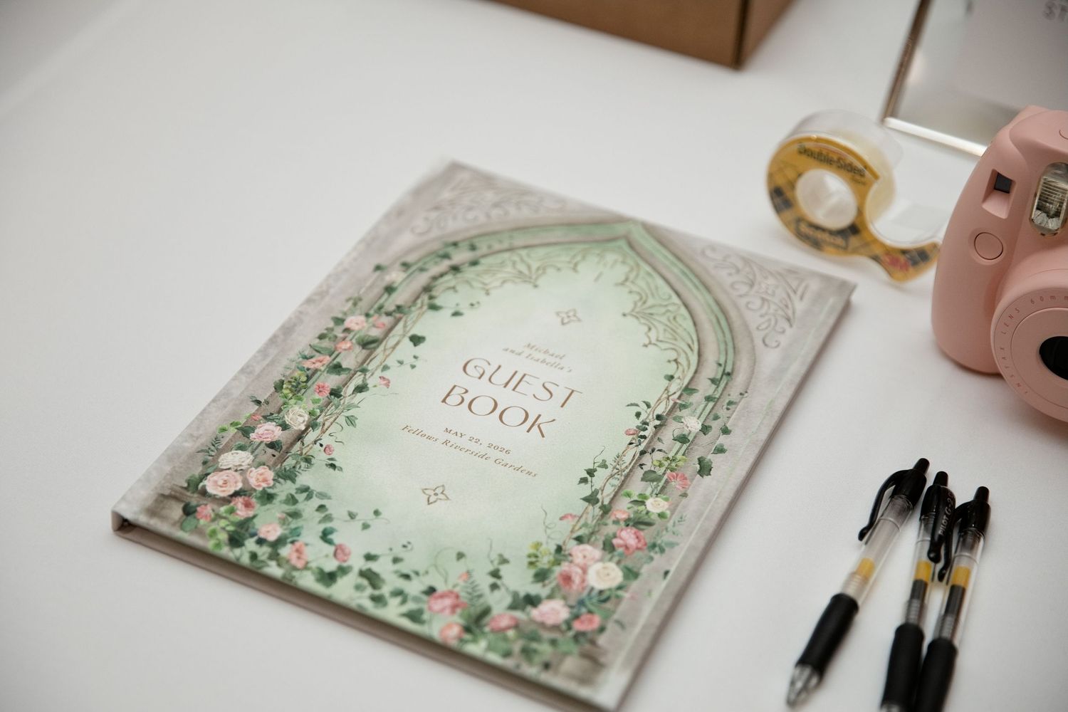 Elegant floral guest book with pink roses on white table beside gold magnifier, pink camera, and black pens.