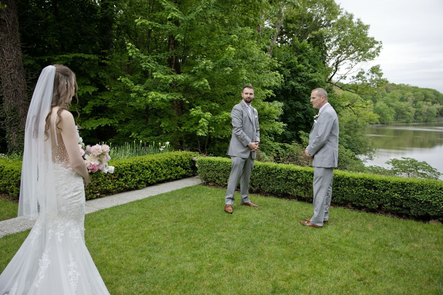 Bride in lace gown and veil walks toward groom and groomsman on green lawn by lake at outdoor wedding.