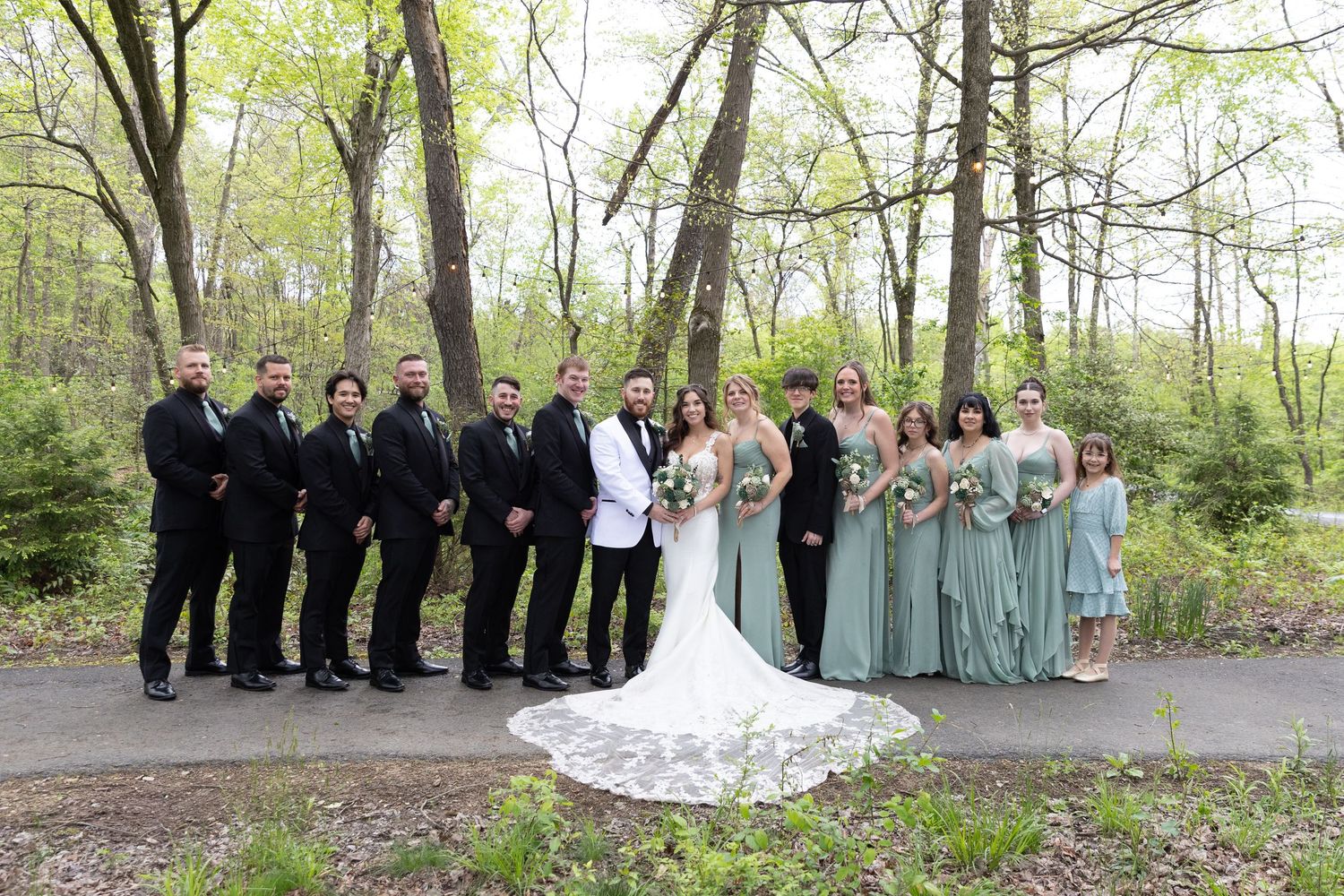 Wedding party poses outdoors in forest setting, bride in lace gown with groomsmen in black and bridesmaids in sage green.