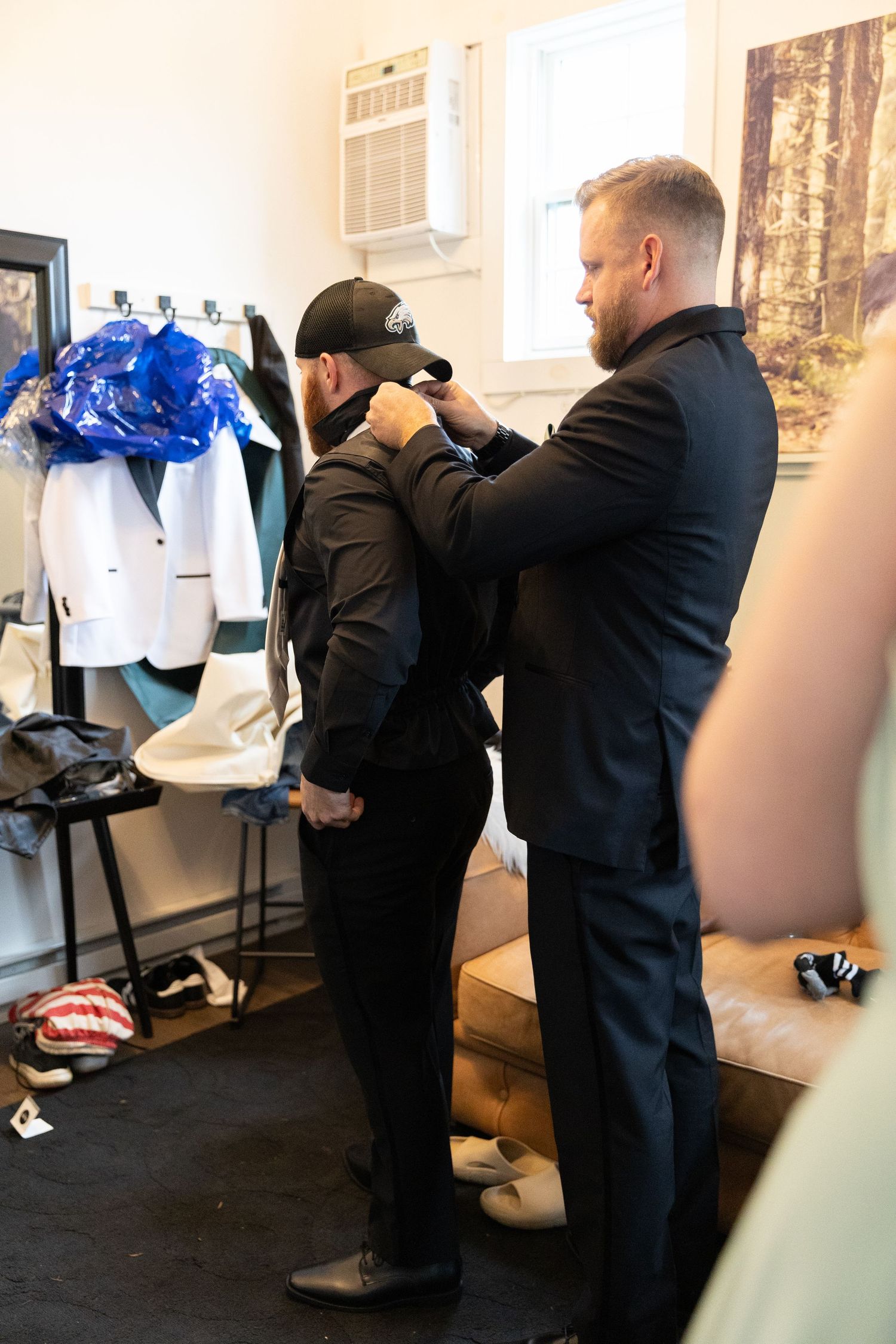 Two men in black suits in a fitting room, one adjusting the other's jacket near a rack of formal wear.