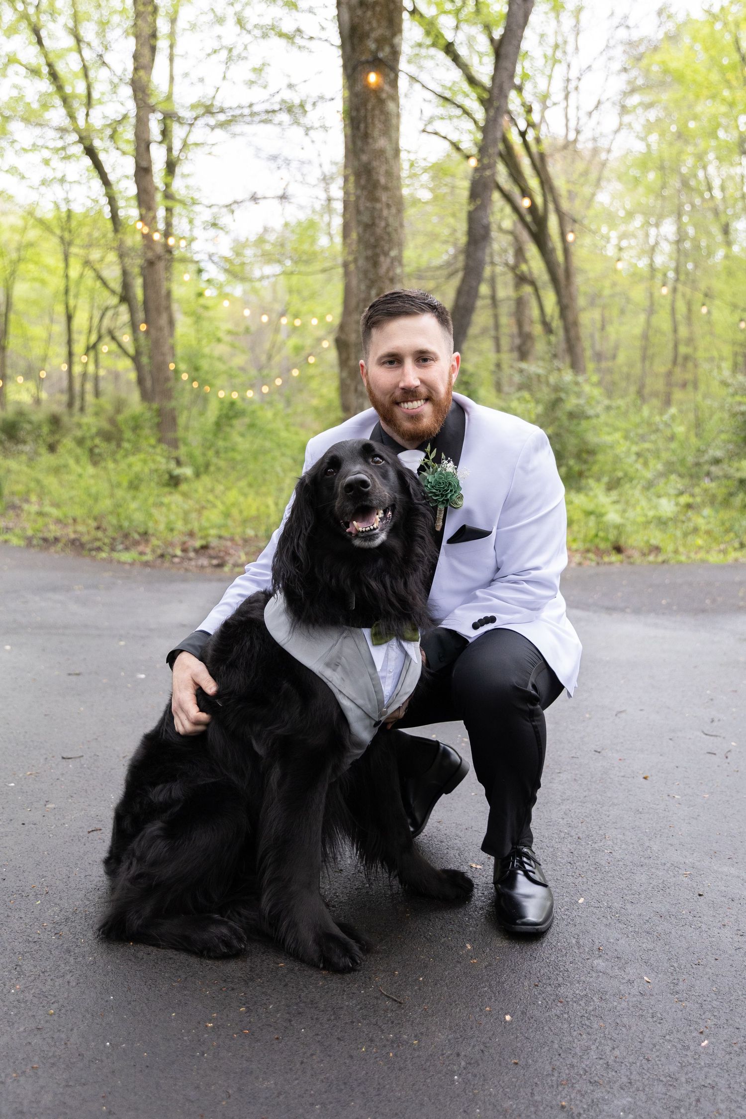 Smiling groom in white tuxedo jacket poses with large black dog wearing matching outfit outdoors.